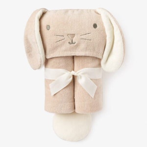 May include: A beige hooded towel with bunny ears and a white ribbon. The towel has embroidered bunny features, including eyes, whiskers, and a nose.