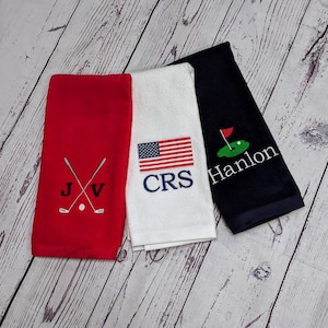 May include: Three golf towels in red, white, and navy blue. The red towel has crossed golf clubs and the initials "J V". The white towel displays an American flag and the initials "CRS". The navy towel features a golf green with a flag and the name "Hanlon".
