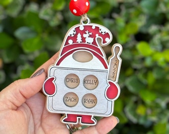Op de afbeelding: Een houten kerstversiering in de vorm van een gnome die een deegroller vasthoudt. De gnome draagt ​​een rode muts met witte sneeuwvlokken en een rendierontwerp. De versiering heeft vier cirkels met de namen Chris, Kelly, Zach en Ryan. De versiering heeft ook het jaar 2023 erop.