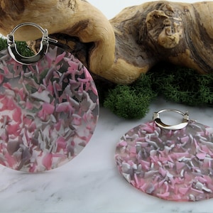 May include: A pair of large, circular earrings with a pink, grey, and white confetti design. They feature silver-coloured hoops. The background includes a piece of driftwood and green moss.