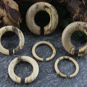 May include: Set of six wooden ear gauges in various sizes. The gauges are made of natural wood with a light brown color and dark brown wood grain patterns.