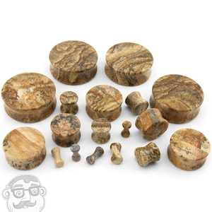 May include: A collection of brown and tan stone ear plugs in various sizes and shapes. The plugs have a natural, marbled appearance, with variations in color and texture. Some are cylindrical, others are flared or hourglass-shaped.