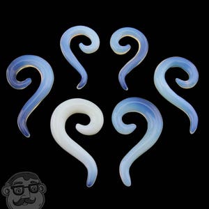 Op de afbeelding: Zes opalescerende glazen oor spiralen in tinten wit en blauw, tegen een zwarte achtergrond. De spiralen hebben een gebogen, langwerpige vorm, met een gladde, gepolijste afwerking. Het ontwerp is symmetrisch en elegant, geschikt voor body modification.