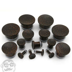 May include: An assortment of dark brown wooden ear plugs. The plugs have a textured surface and a flared design with a clear, rubber-like ring. The collection includes single and double-flare styles. Sizes vary, suitable for stretched ear piercings. Measurements are in centimetres.