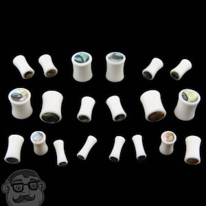 May include: A collection of white acrylic ear gauges with abalone shell inlay. The gauges are various sizes and are displayed on a black background.
