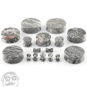 May include: A collection of gray and white marbled stone ear plugs and gauges in various sizes and shapes. The plugs are round, oval, and hourglass-shaped. The stone has a natural, swirled pattern. The gauges are small and bone-shaped.