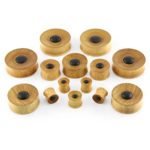 May include: A collection of wooden ear plugs in various sizes. Each plug features a light brown, circular wooden base with a dark, circular center. The plugs are designed with a flared shape, and are displayed against a white background.