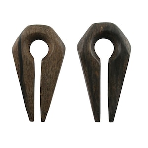 May include: Two dark brown wooden ear gauges with a geometric design. Each gauge has a rounded top with a central hole and a split, elongated bottom. The wood grain is visible, adding texture to the plugs.