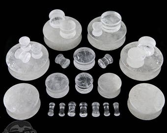 May include: A set of clear quartz ear gauges in various sizes, displayed on white quartz bases. The gauges are smooth and polished, with a natural crystal look.