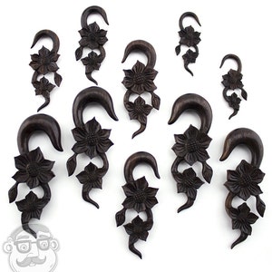 Könnte beinhalten: Ein Set aus neun dunkelbraunen Holz-Ohrenstreckern mit floralem Design. Jeder Strecker hat einen gebogenen Haken und drei geschnitzte Blumen.