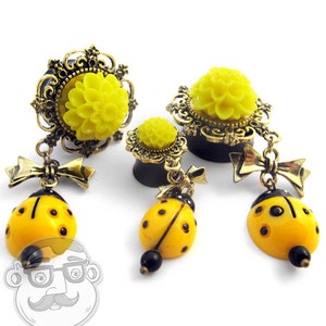 May include: Ear gauges with yellow flower and ladybug charms. The gauges have a black base with a yellow flower design and a gold filigree frame. The ladybug charms are yellow with black spots and a black bead.