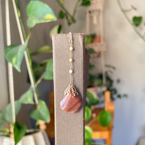 Könnte beinhalten: Ein Edelsteinanhänger mit einer goldfarbenen Filigran-Kappe und einer zarten Kette aus kleinen weißen Perlen. Der Anhänger hat eine rotbraune Farbe mit helleren Streifen. Der Hintergrund umfasst grüne Pflanzen und eine Holzstruktur.