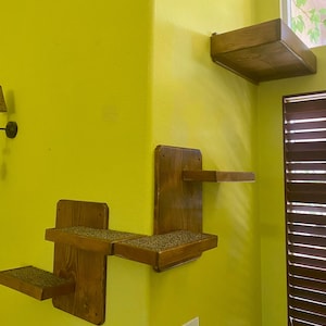 May include: A set of three wooden cat shelves mounted on a yellow wall. The shelves are in a corner and are designed to be used by cats to climb and play. The shelves are made of natural wood and have a simple, modern design.