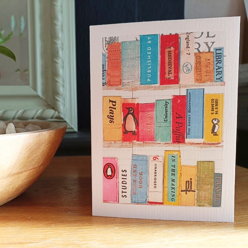 May include: A greeting card featuring a colorful illustration of bookshelves filled with vintage books. The card includes titles such as "Plays," "Studies," and "Medieval." A penguin illustration is visible on one of the book spines.