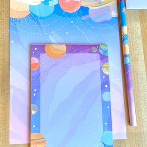 Può includere: Due fogli di carta da lettere con un design a tema spaziale. Il foglio superiore presenta uno sfondo blu chiaro con pianeti e stelle. Il foglio inferiore ha uno sfondo viola con una cornice bianca e pianeti attorno al bordo.