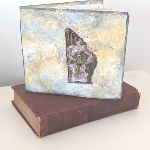 May include: A handmade journal with a textured, painted cover in shades of blue, beige, and gray, adorned with a wooden accent, lace, and a decorative key. The journal rests on a vintage, burgundy-colored book.