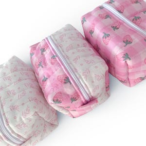 May include: Three quilted cosmetic bags in pink and white. The bags have a zip closure and are decorated with strawberry and floral patterns. The bags are arranged diagonally against a white background.