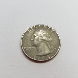 May include: A silver US quarter dollar coin with the year 1984 inscribed on the bottom. The coin features a profile of George Washington with the words "LIBERTY" above and "IN GOD WE TRUST" below.
