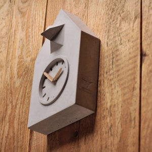 Könnte beinhalten: Eine graue Beton-Kuckucksuhr mit weißem Zifferblatt und Holzzeigern. Die Uhr ist in Form eines Hauses mit einem kleinen Dach.