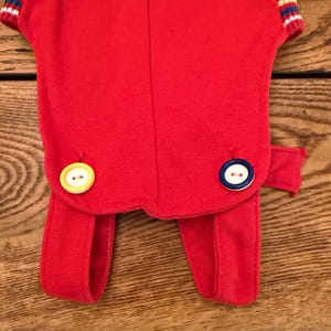 Puede incluir: Un atuendo rojo con una parte superior de manga corta y pantalones adjuntos. La parte superior tiene pu&ntilde;os a rayas en blanco, azul y rojo. El atuendo presenta dos botones grandes, uno amarillo y otro azul y blanco.