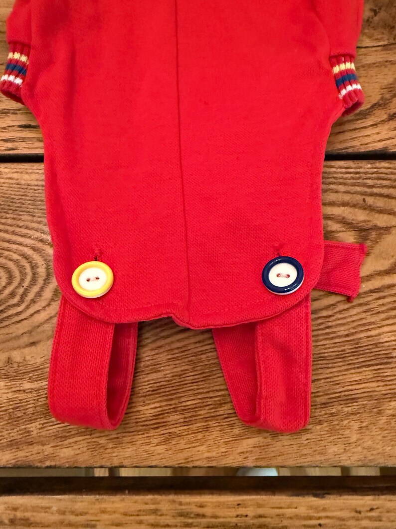 Puede incluir: Un atuendo rojo con una parte superior de manga corta y pantalones adjuntos. La parte superior tiene pu&ntilde;os a rayas en blanco, azul y rojo. El atuendo presenta dos botones grandes, uno amarillo y otro azul y blanco.