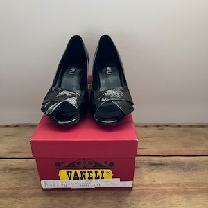 May include: A pair of black patent leather peep-toe heels with a bow detail. The shoes are in a box with the brand name "Vanell" and the model name "Lillie Black Krinkpat" printed on it. The size is 7M, 18883, U14.