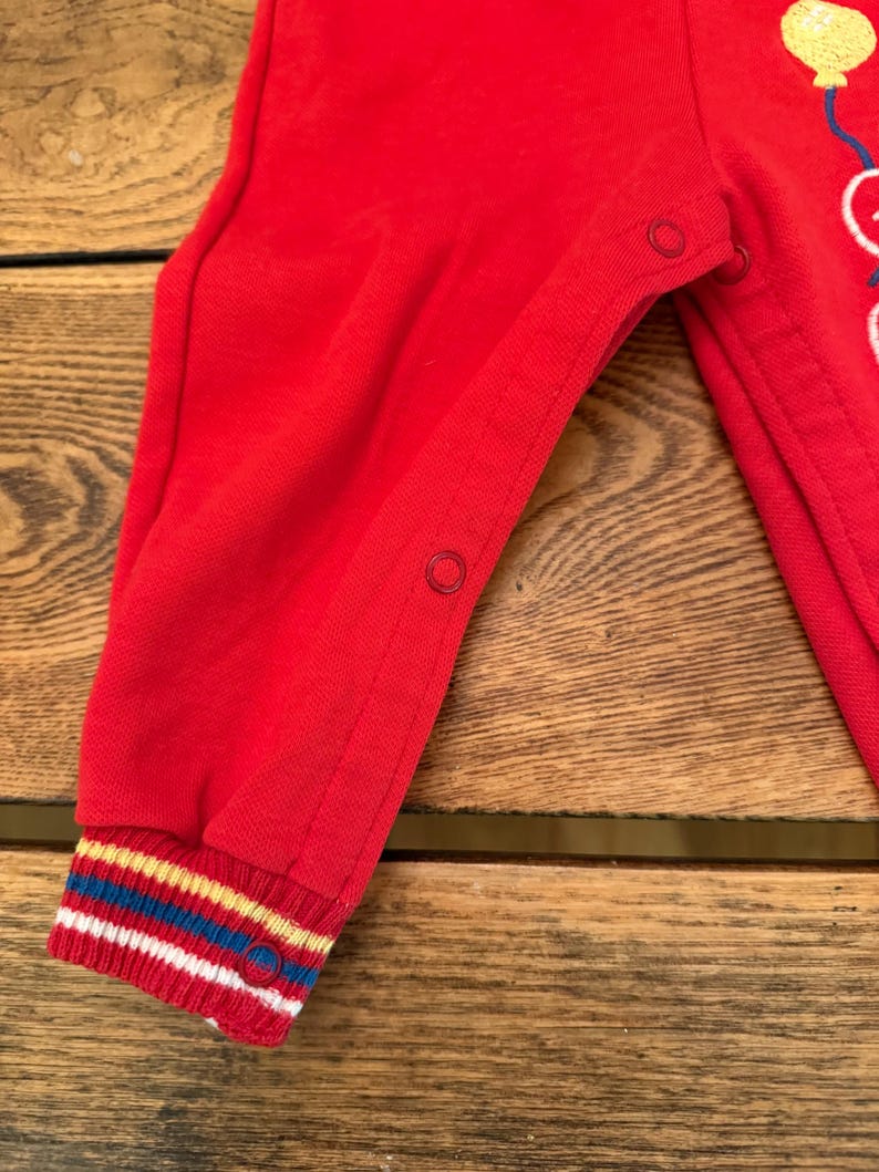 Puede incluir: Mono rojo para beb&eacute; con pu&ntilde;o a rayas de colores. El mono tiene cierres de presi&oacute;n y un gr&aacute;fico de globos. El pu&ntilde;o presenta rayas blancas, azules, amarillas y rojas.