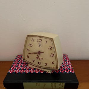 May include: A white vintage alarm clock with black numbers and hands. The clock has a square face and a rounded rectangular body. The clock is sitting on a book with a red and blue patterned cover. The book has the words "Best Sellers" printed on the spine.
