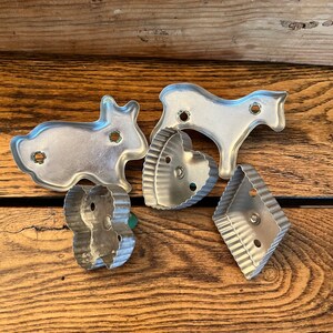 May include: Five vintage metal cookie cutters in various shapes, including a rabbit, a horse, a heart, and two different diamond shapes. The cutters are silver in color and have a slightly worn look.