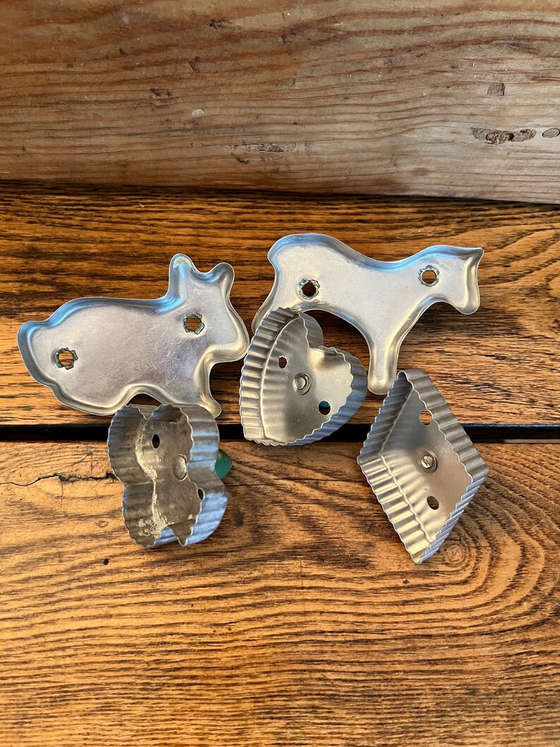 May include: Five vintage metal cookie cutters in various shapes, including a rabbit, a horse, a heart, and two different diamond shapes. The cutters are silver in color and have a slightly worn look.