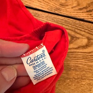 Puede incluir: Primer plano de una prenda roja brillante con una etiqueta blanca. La etiqueta dice "Carter's MADE IN U.S.A. 18 MESES 10.4-11.8 kg." La prenda est&aacute; hecha de 50% poli&eacute;ster y 50% algod&oacute;n. La etiqueta tambi&eacute;n incluye instrucciones de lavado.