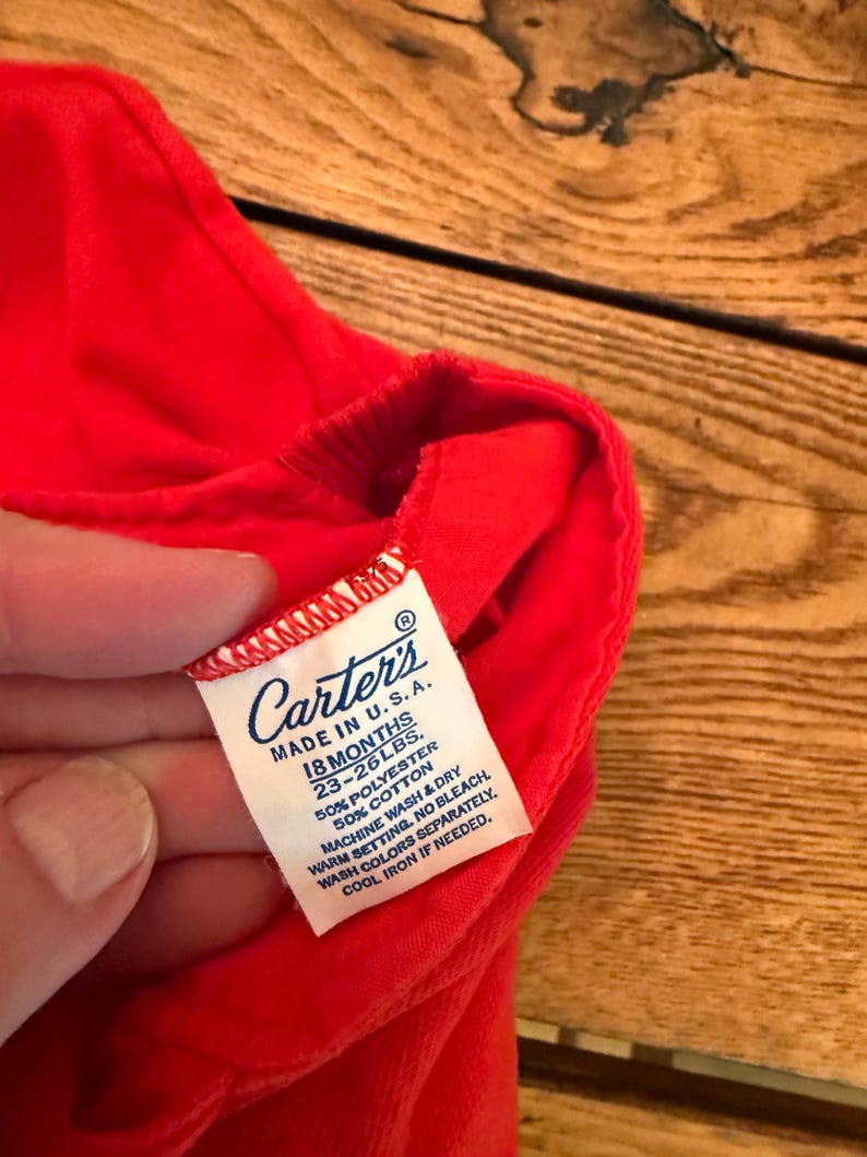 Puede incluir: Primer plano de una prenda roja brillante con una etiqueta blanca. La etiqueta dice "Carter's MADE IN U.S.A. 18 MESES 10.4-11.8 kg." La prenda est&aacute; hecha de 50% poli&eacute;ster y 50% algod&oacute;n. La etiqueta tambi&eacute;n incluye instrucciones de lavado.
