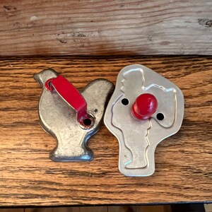 May include: Two vintage metal cookie cutters, one shaped like a bird and the other shaped like a person. Both cutters have red plastic handles.