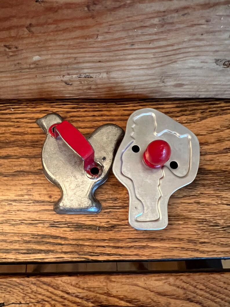 May include: Two vintage metal cookie cutters, one shaped like a bird and the other shaped like a person. Both cutters have red plastic handles.