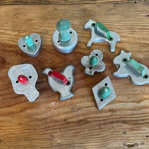 May include: A set of nine vintage metal cookie cutters with wooden handles. The cutters are shaped like a heart, a horse, a chicken, a diamond, a clover, a bull, a man, and two round cutters.