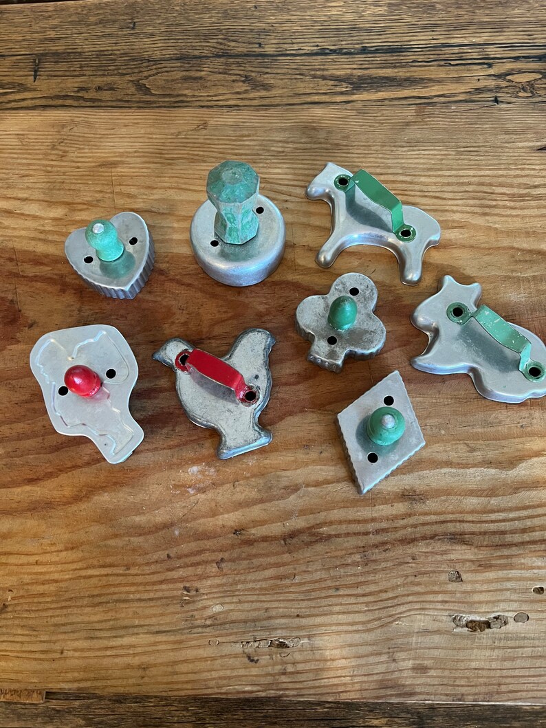 May include: A set of nine vintage metal cookie cutters with wooden handles. The cutters are shaped like a heart, a horse, a chicken, a diamond, a clover, a bull, a man, and two round cutters.