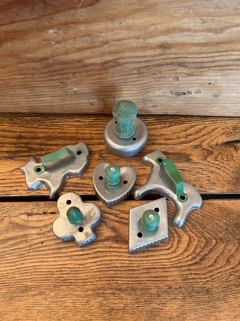 May include: A set of six vintage metal cookie cutters with green wooden handles. The cutters are shaped like a heart, a diamond, a clover, a horse, a bunny, and a round cutter.