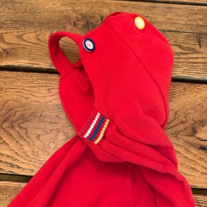 Puede incluir: Una prenda con capucha de color rojo brillante con un lazo y dos botones, amarillo y azul. El pu&ntilde;o de la manga tiene un dise&ntilde;o a rayas con rayas blancas, azules y amarillas. La prenda est&aacute; hecha de una tela de punto suave.