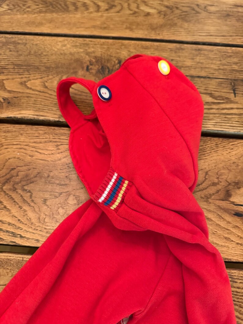 Puede incluir: Una prenda con capucha de color rojo brillante con un lazo y dos botones, amarillo y azul. El pu&ntilde;o de la manga tiene un dise&ntilde;o a rayas con rayas blancas, azules y amarillas. La prenda est&aacute; hecha de una tela de punto suave.