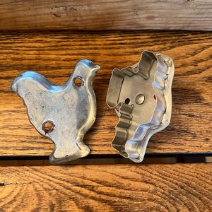 May include: Two vintage metal cookie cutters, one shaped like a chicken and the other shaped like a Christmas tree. The cutters are silver and have a worn, antique look.