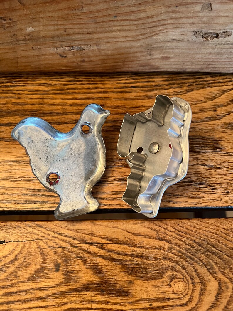 May include: Two vintage metal cookie cutters, one shaped like a chicken and the other shaped like a Christmas tree. The cutters are silver and have a worn, antique look.