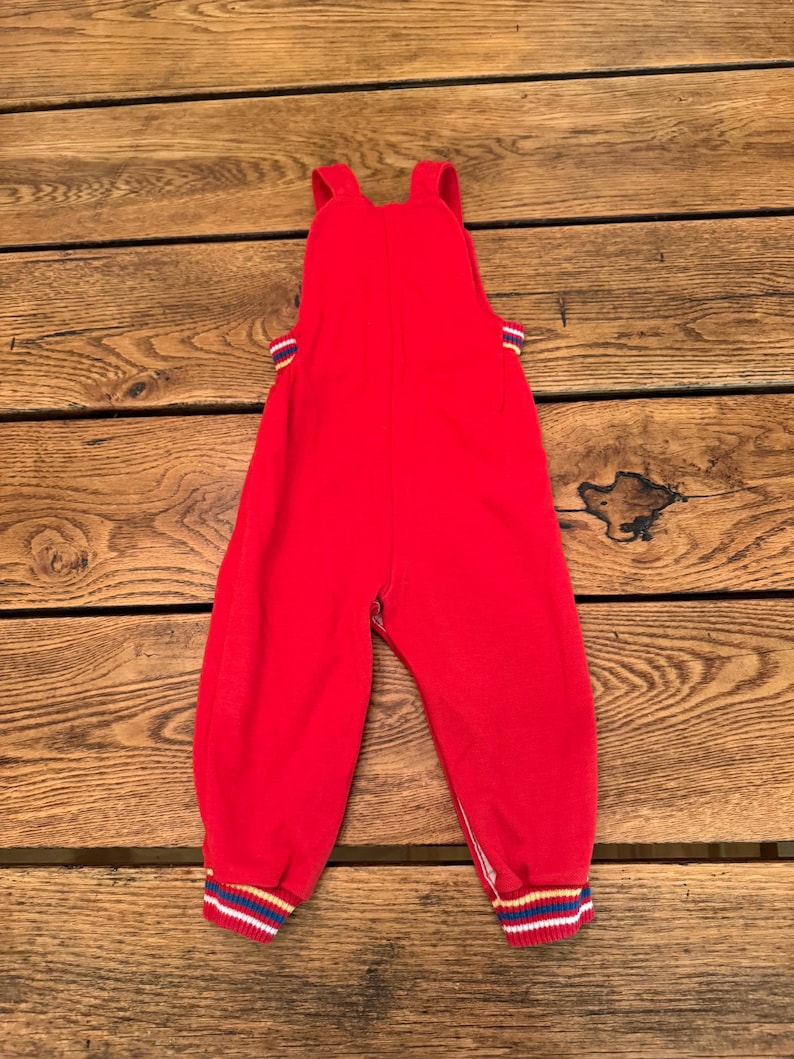 Puede incluir: Mono rojo para beb&eacute; con tirantes. Los pu&ntilde;os y los lados del mono tienen un estampado a rayas en azul, amarillo y blanco. El mono est&aacute; hecho de un material suave.