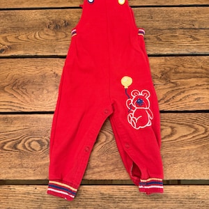 Puede incluir: Mono rojo para beb&eacute; con un gr&aacute;fico de oso y globo. El mono tiene cierres de bot&oacute;n en las correas y las piernas. Los pu&ntilde;os presentan rayas azules, amarillas y rojas. El mono est&aacute; hecho de un material suave.