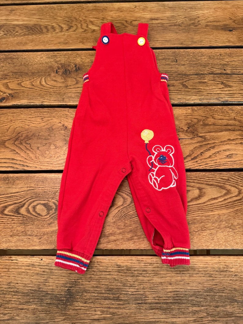 Puede incluir: Mono rojo para beb&eacute; con un gr&aacute;fico de oso y globo. El mono tiene cierres de bot&oacute;n en las correas y las piernas. Los pu&ntilde;os presentan rayas azules, amarillas y rojas. El mono est&aacute; hecho de un material suave.