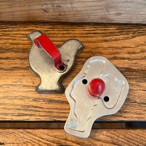 May include: Two vintage metal cookie cutters, one shaped like a turkey with a red handle and the other shaped like a snowman with a red nose.