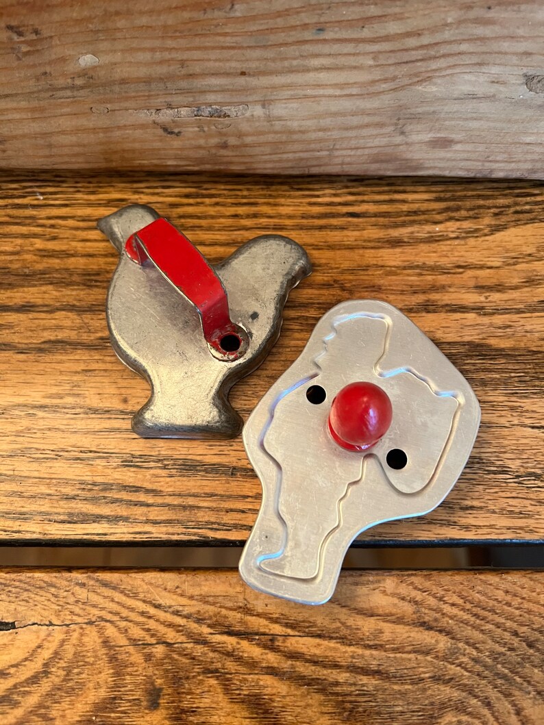May include: Two vintage metal cookie cutters, one shaped like a turkey with a red handle and the other shaped like a snowman with a red nose.