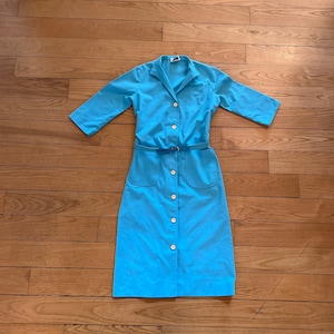 May include: A light blue dress with a collared neckline, three-quarter sleeves, and a button-down front. The dress features a matching belt and two front pockets. The buttons are a light gold color.