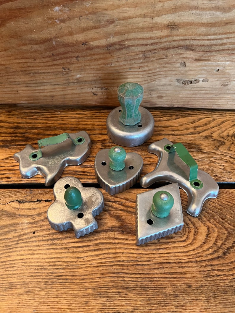 May include: A set of six vintage metal cookie cutters with green wooden handles. The cutters are shaped like a heart, a diamond, a club, a dog, a cat, and a round cutter.