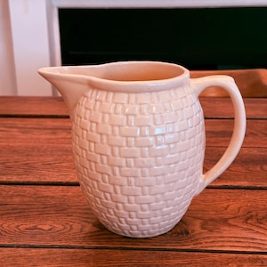 May include: A cream-colored ceramic pitcher with a basketweave pattern. The pitcher has a spout and a curved handle. It is sitting on a wooden surface.