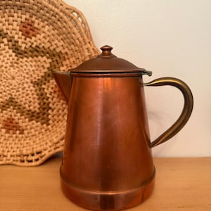 May include: A copper coffee pot with a lid and a brass handle. The pot is sitting on a wooden surface. A woven basket is in the background.